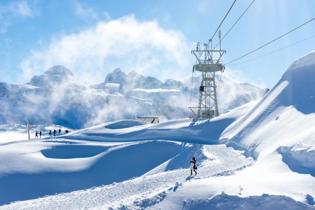 Eine Frau geht auf einem verschneiten Pfad in den Bergen entlang, im Hintergrund ein Liftmast.