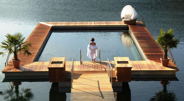 Eine Frau in einem weißen Bademantel steht am Rand eines Pools auf einer Holzplattform im Wasser.