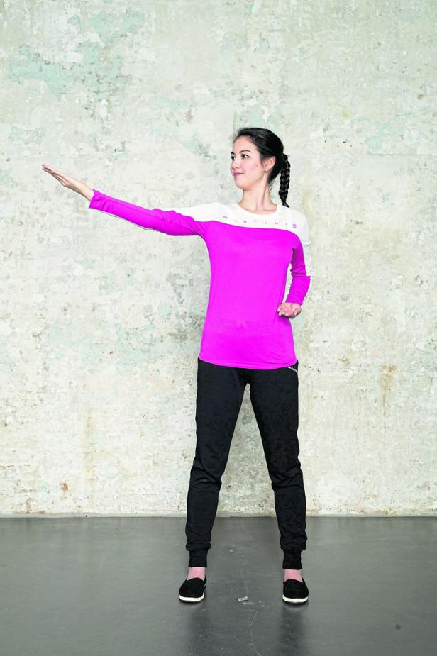 Eine Frau in Sportkleidung steht vor einer Wand und streckt den Arm aus.