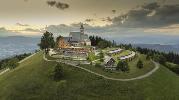 Ein Hotel und eine Kirche stehen auf einem grünen Hügel mit Blick auf eine Berglandschaft.