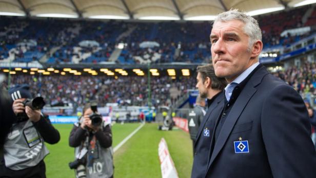 Ein Mann im Anzug steht am Spielfeldrand vor einer Zuschauertribüne.