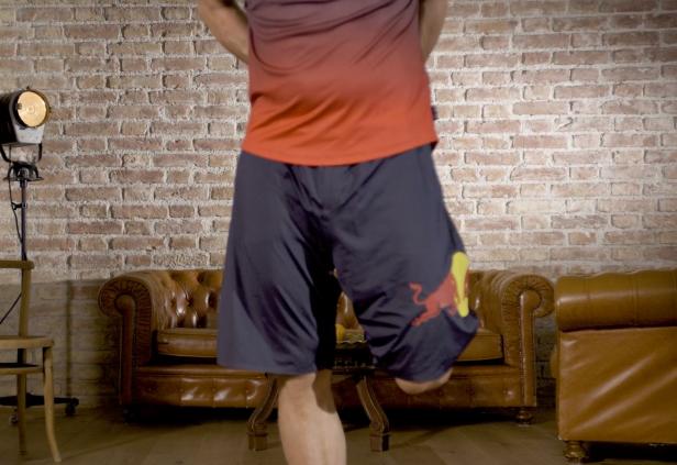 Ein Mann in Sportkleidung mit Red Bull Logo macht Übungen in einem Wohnzimmer.