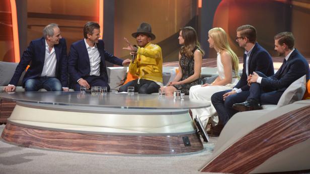 Mehrere Personen sitzen auf einer Couch während einer Fernsehsendung, darunter Pharrell Williams.