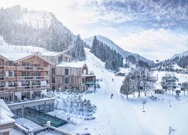 Winterlandschaft mit schneebedeckten Bergen, einem Hotel und Skifahrern auf der Piste.