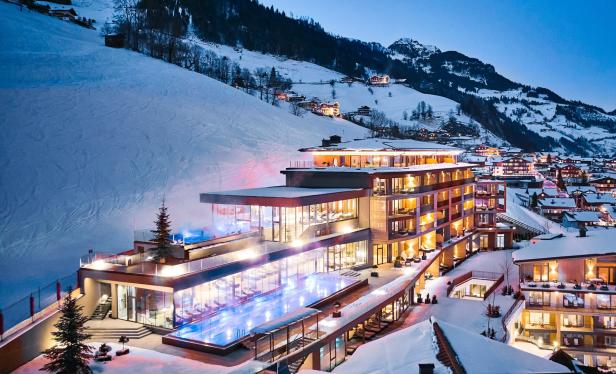 Ein modernes Hotel mit beleuchteten Pools liegt in einer verschneiten Berglandschaft.
