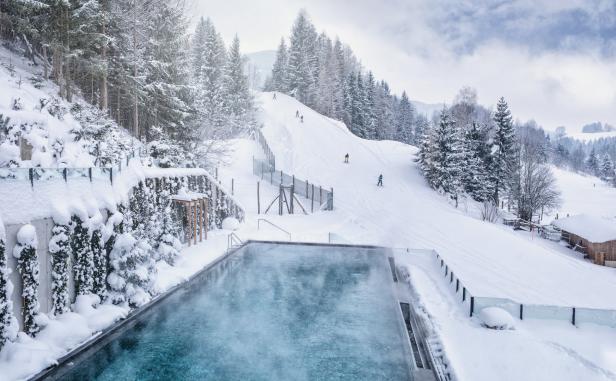Ein beheizter Außenpool mit Blick auf eine schneebedeckte Skipiste mit Skifahrern.