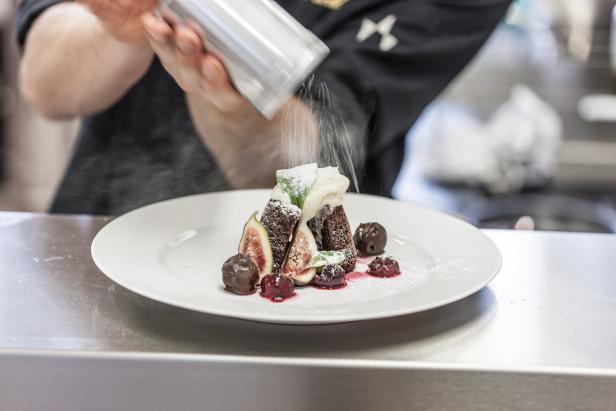 Ein Koch bestreut ein Dessert mit Feigen, Schokoladenkugeln und Puderzucker.