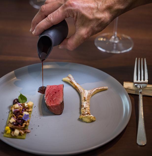 Ein Teller mit einem Stück Fleisch, einer Geweih-Dekoration und einer grünen Beilage, übergossen mit Sauce.