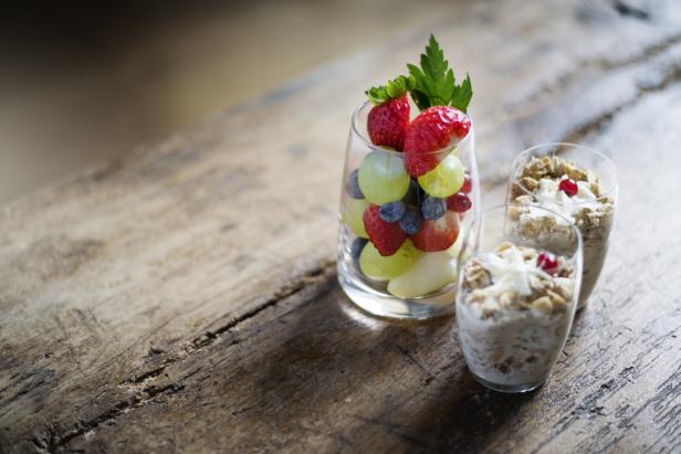 Ein Glas mit Erdbeeren, Blaubeeren und Weintrauben neben zwei Gläsern Müsli auf einem Holztisch.