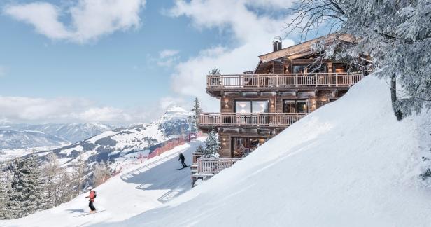 Ein luxuriöses Holzchalet steht an einem verschneiten Hang, während Skifahrer vorbeifahren.