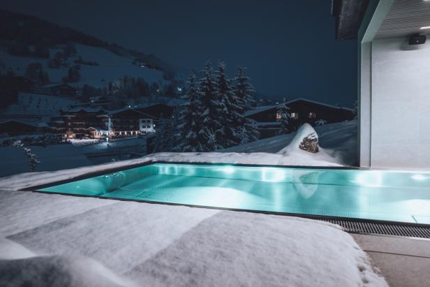 Ein beleuchteter Pool ist von Schnee umgeben, im Hintergrund ein verschneites Dorf bei Nacht.