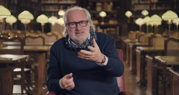 Ein Mann mit Brille und Schal gestikuliert in einer Bibliothek.