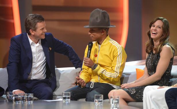 Pharrell Williams, ein Mann im Anzug und Hilary Swank sitzen auf einer Couch.