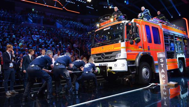 Mehrere Männer ziehen ein Feuerwehrfahrzeug vor Publikum.