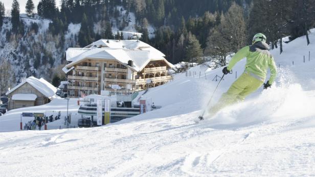 Ein Skifahrer fährt eine schneebedeckte Piste vor einem Hotel hinunter.