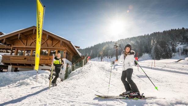 Zwei Frauen stehen auf einer Skipiste vor einem hölzernen Gebäude.
