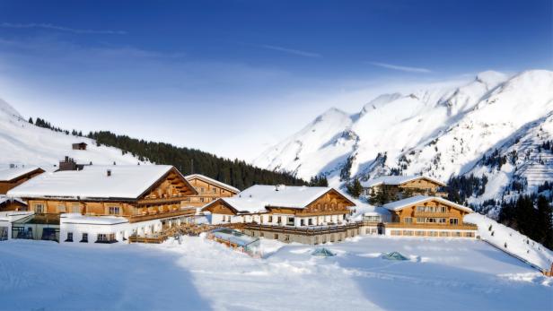 Mehrere Holzhäuser sind im Winter mit Schnee bedeckt, im Hintergrund schneebedeckte Berge.