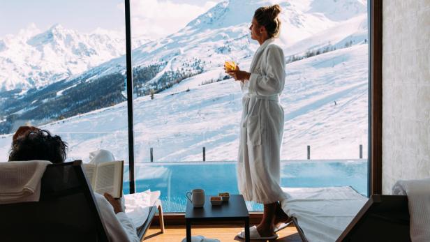 Eine Frau im Bademantel genießt einen Drink mit Blick auf die schneebedeckten Berge.