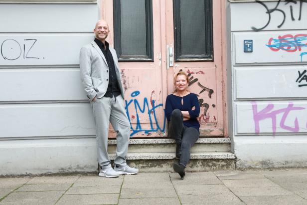 Ein Mann im Anzug steht neben einer Frau, die vor einer mit Graffiti besprühten Tür sitzt.