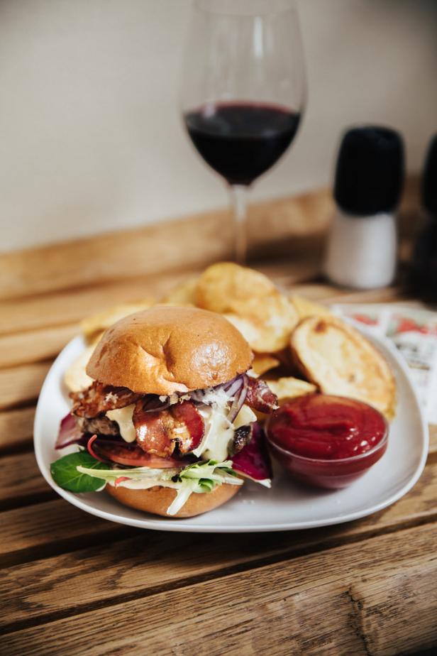 Ein Burger mit Speck, Käse und Pommes auf einem Teller mit einem Glas Rotwein im Hintergrund.