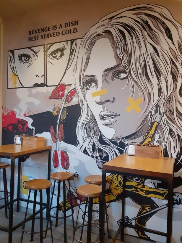 Ein Wandgemälde mit dem Zitat „Revenge is a dish best served cold“ und Comic-Elementen in einem Restaurant.