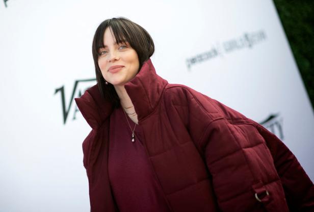 Billie Eilish trägt eine weinrote Jacke und steht vor einem weißen Hintergrund.