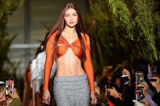 Gigi Hadid läuft in einem orangefarbenen Oberteil und einem silbernen Rock über einen Laufsteg.