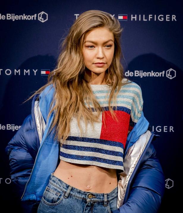 Gigi Hadid trägt ein bauchfreies Top und eine blaue Jacke vor einem Tommy Hilfiger Hintergrund.