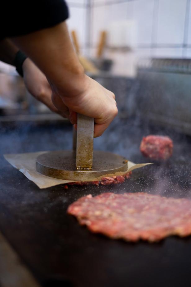 Eine Person presst mit einem Burger-Pressegerät Hackfleisch auf einer heißen Grillplatte.