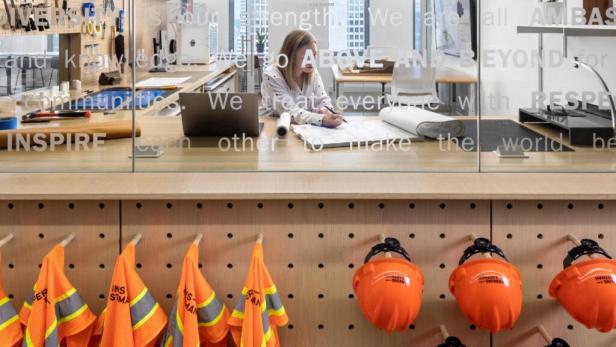 Eine Frau arbeitet an einem Bauplan in einem Büro, im Vordergrund hängen Sicherheitswesten und Helme.