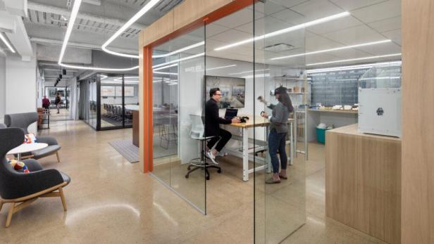 In einem modernen Büro arbeiten zwei Personen mit einem Laptop und einer VR-Brille in einem gläsernen Raum.