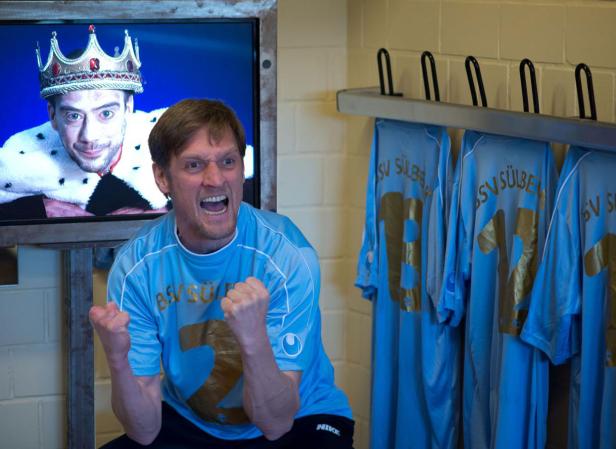 Ein Mann im Trikot des SV Sülbeck jubelt vor einem Fernseher, auf dem ein Mann mit Krone zu sehen ist.