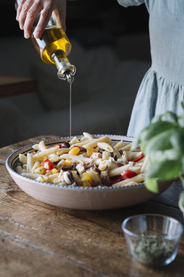 Eine Person beträufelt einen Teller Penne-Nudelsalat mit Olivenöl.