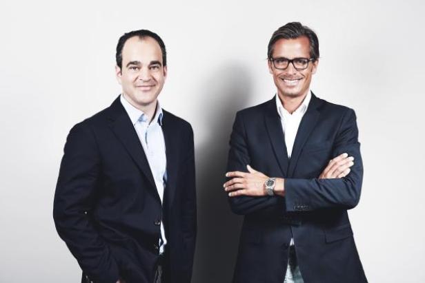 Zwei Männer in Anzügen stehen vor einem hellen Hintergrund.