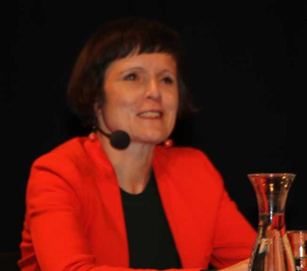 Eine Frau mit Mikrofon und rotem Blazer lächelt.