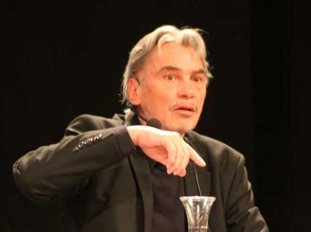 Ein Mann mit grauem Haar gestikuliert während einer Rede vor einem dunklen Hintergrund.