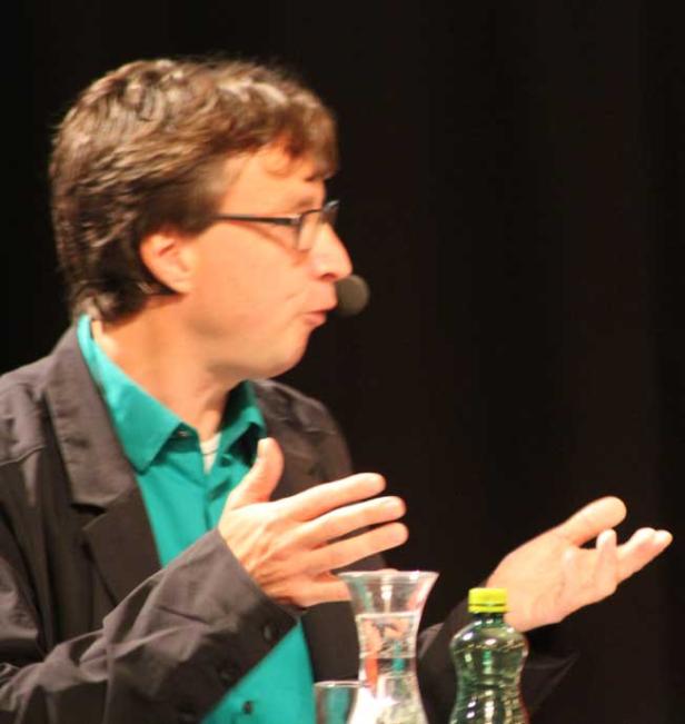 Ein Mann mit Brille gestikuliert während eines Vortrags mit Mikrofon.