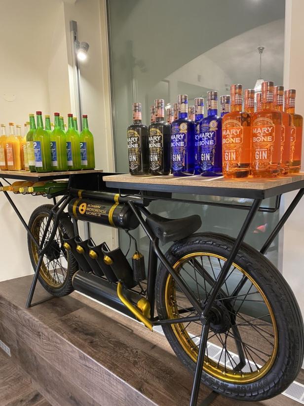Ein Regal in Form eines Motorrads präsentiert Flaschen von „Mary Bone Gin“ in verschiedenen Farben.