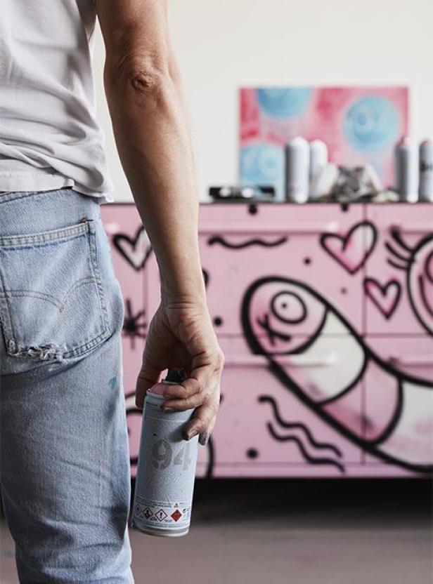 Eine Person hält eine Sprühdose vor eine rosa Kommode mit Graffiti.