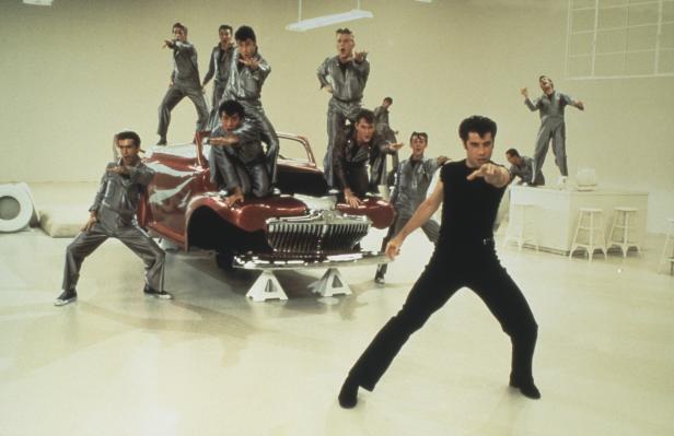 John Travolta und andere Darsteller tanzen um ein Auto im Film „Grease“.