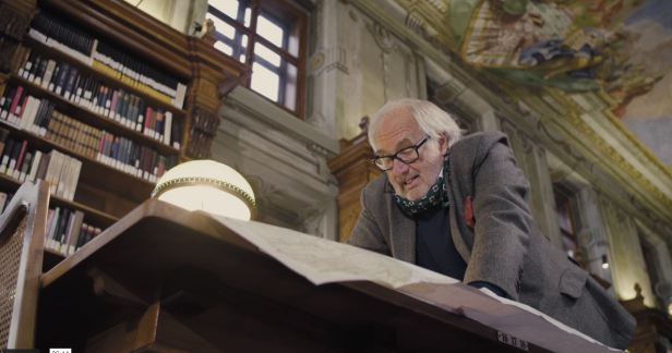Ein älterer Mann mit Brille betrachtet eine Karte in einer Bibliothek.