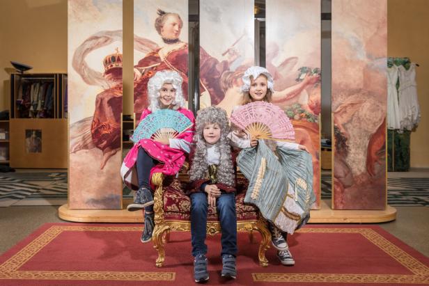 Drei Kinder in historischen Kostümen und Perücken sitzen auf einem goldenen Stuhl und halten Fächer.