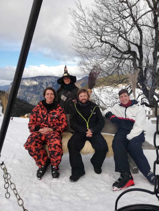 Eine Gruppe von vier Personen sitzt auf einer Bank im Schnee, eine davon mit Hexenhut und Besen.