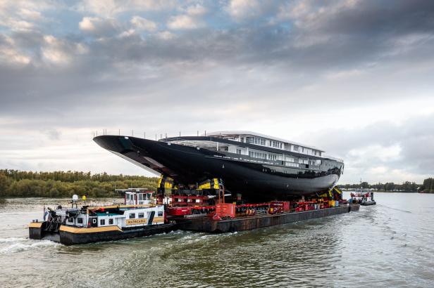 Eine große, schwarze Yacht wird auf einem Ponton von Schleppern einen Fluss entlang transportiert.