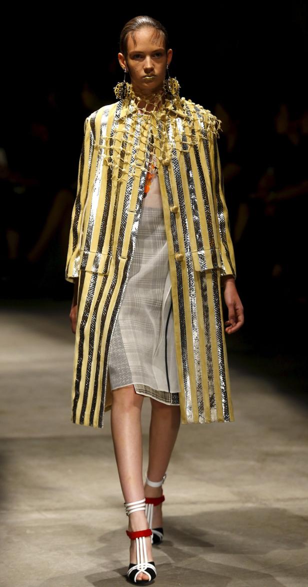 Ein Model präsentiert einen gestreiften Mantel und auffälligen Schmuck auf dem Laufsteg.