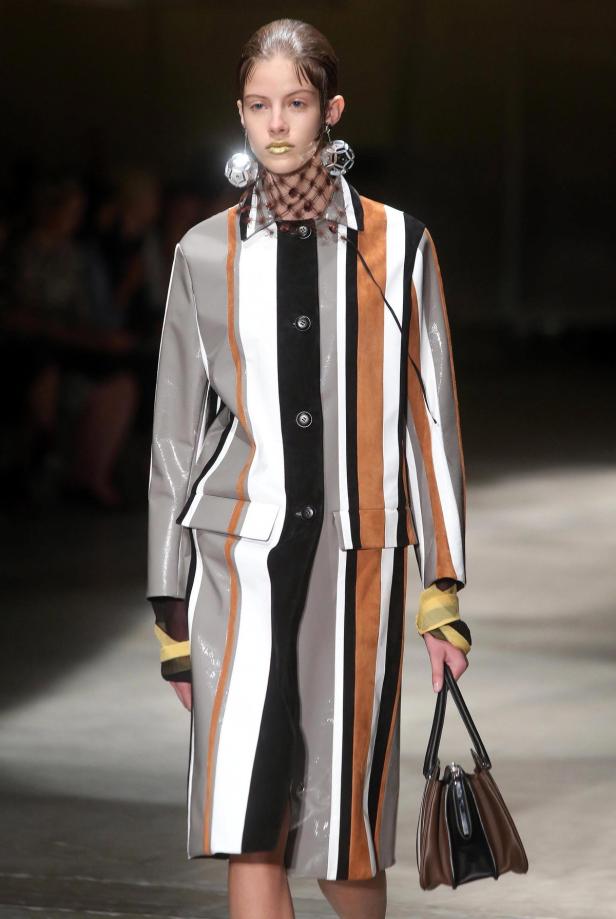 Ein Model präsentiert einen gestreiften Mantel, auffällige Ohrringe und goldfarbenen Lippenstift auf dem Laufsteg.