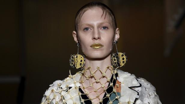 Ein Model mit goldenen Lippen und auffälligen Ohrringen auf dem Laufsteg.