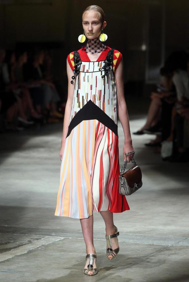 Ein Model präsentiert ein gestreiftes Kleid, auffällige Ohrringe und eine Handtasche auf dem Laufsteg.