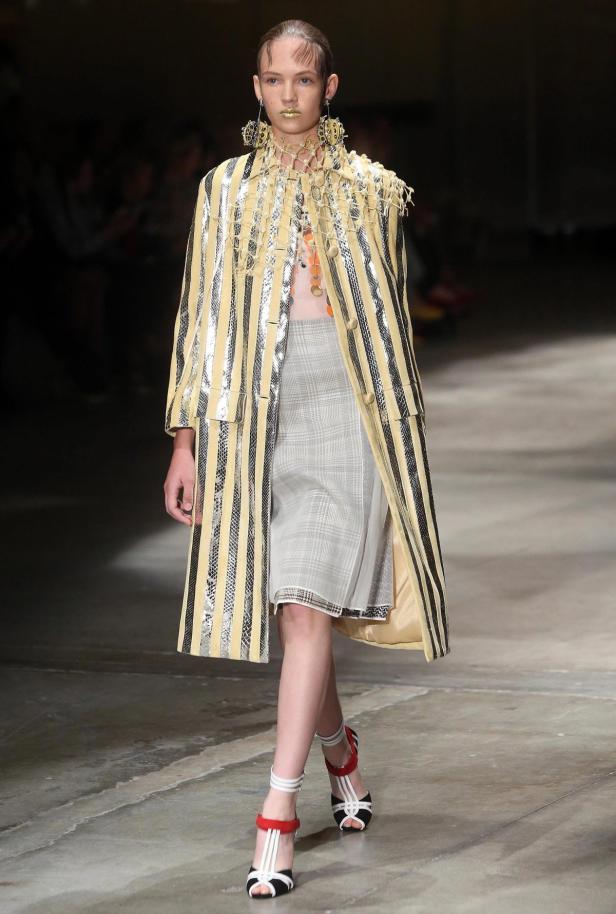 Ein Model präsentiert einen gestreiften Mantel, einen karierten Rock und auffälligen Schmuck auf dem Laufsteg.