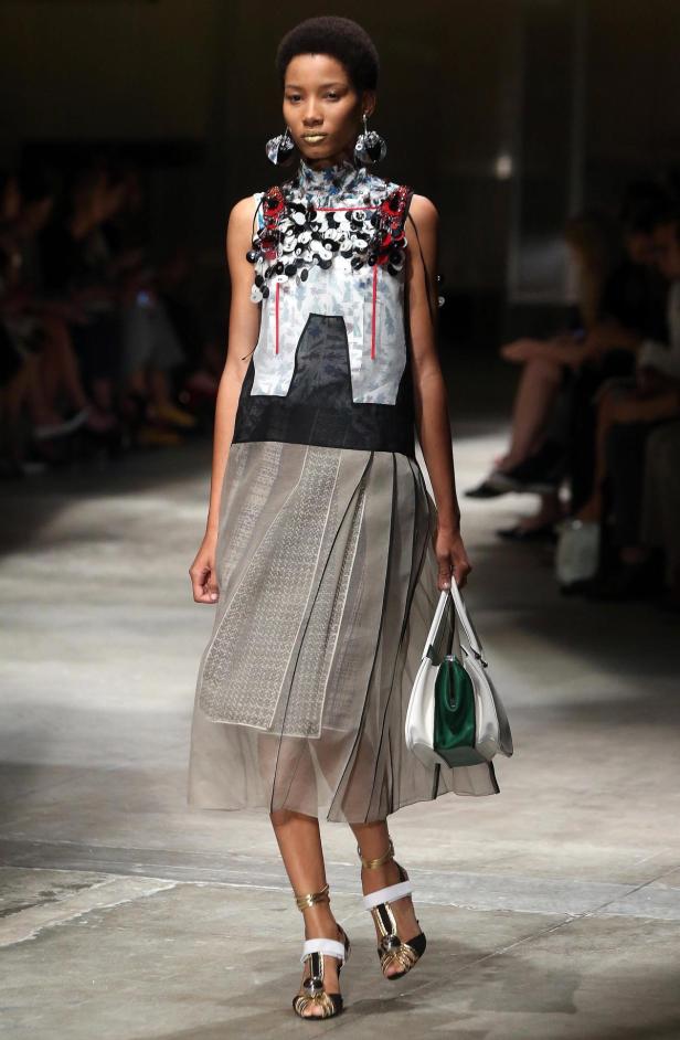 Eine Frau präsentiert ein ärmelloses Kleid mit Applikationen und eine Handtasche auf dem Laufsteg.